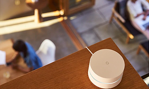Google Wifi-enhet placerad högt upp på en trähylla. Nedanför syns en person som sitter vid ett bord.