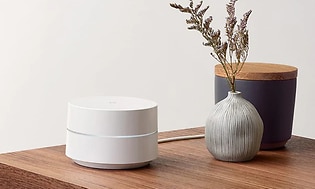Vit rund Google Wifi-enhet placerad på ett träbord bredvid en vas och andra dekorationer. 
