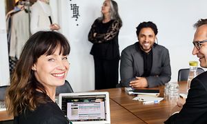 En kvinde og to mænd sidder smilende rundt et bord i et mødelokale og arbejder på en bærbar computer