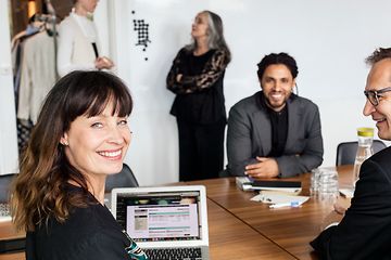 En kvinde og to mænd sidder smilende rundt et bord i et mødelokale og arbejder på en bærbar computer