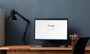 Svart datorskärm på ett skrivbord i trä med svart skrivbordsstol och lampa, på skörmen syns google. 