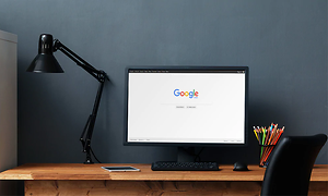 Svart datorskärm på ett skrivbord i trä med svart skrivbordsstol och lampa, på skörmen syns google.