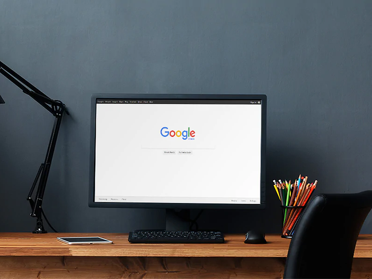 Svart datorskärm på ett skrivbord i trä med svart skrivbordsstol och lampa, på skörmen syns google.