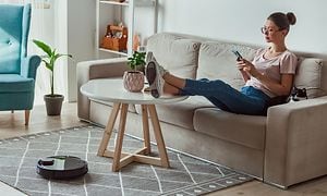 Robotdammsugare på golvet och en kvinna med glasögon och hårknut i en soffa med fötterna på bordet som tittar på sin smartphone