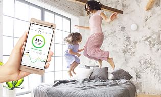 Mamma som hoppar i sängen med sin dotter i bakgrunden och en Airthings-app på en smartphone i förgrunden
