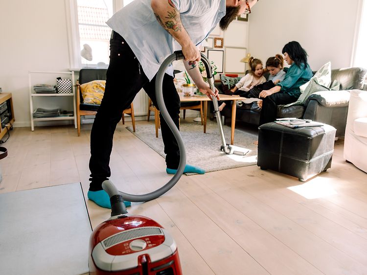 Man som dammsuger medan fru och barn sitter i soffan bakom honom