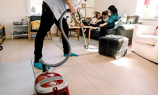 Man som dammsuger medan fru och barn sitter i soffan bakom honom