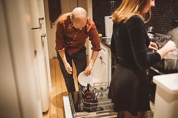 En man och kvinna i ett kök. Man som sätter in disk i diskmaskinen och en kvinna som lagar mat