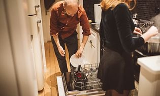 En man och kvinna i ett kök. Man som sätter in disk i diskmaskinen och en kvinna som lagar mat