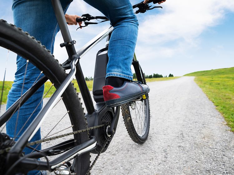 Person i jeans cyklar på en elcykel på en väg med grönt gräs på båda sidor och blå himmel.