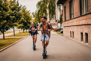 Man och kvinna i full fart på en väg på varsin elsparkcykel. 