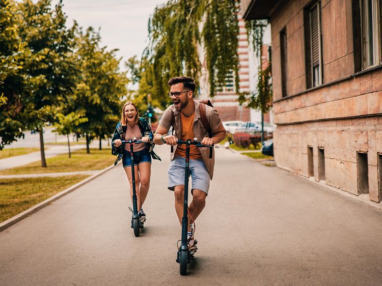 Man och kvinna i full fart på en väg på varsin elsparkcykel. 