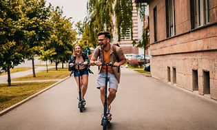 Man och kvinna i full fart på en väg på varsin elsparkcykel. 