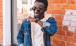 Man i jeansjacka och solglasögon pratar med någon via en smartklocka på handleden.