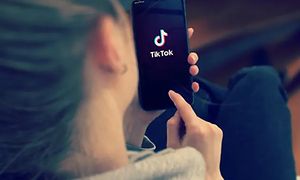 En kvinna håller i en mobiltelefon med en TikTok logga på skärmen. Bild tagen bakifrån, över axeln.