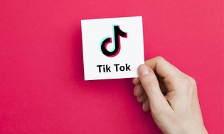 En hand håller i ett vitt kort med TikTok logga på, mörkrosa bakgrund.