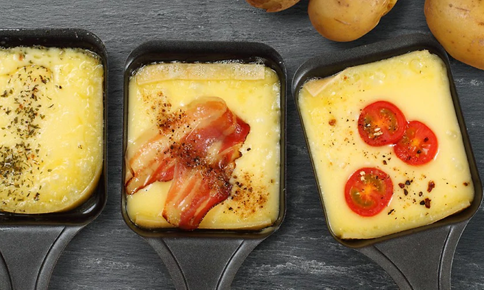 Tre Raclette-pannor med ost, några tomater i den till höger, bacon i den i mitten och kryddor på den till vänster. 