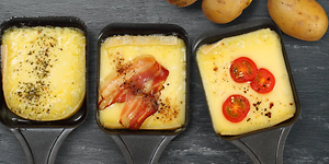 Tre Raclette-pannor med ost, några tomater i den till höger, bacon i den i mitten och kryddor på den till vänster. 