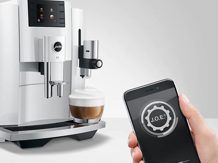 Kaffemaskin från Jura med en färdig kaffe och en hand som håller fram en mobil med texten J.O.E. på skärmen. 