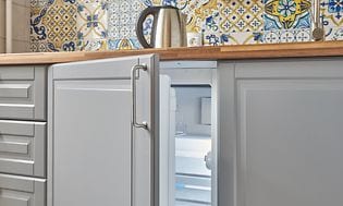 Litet integrerat kylskåp under en köksbänk i trä, grå köksluckor och kakel med mönster på. 