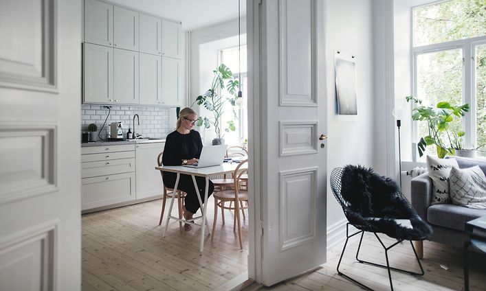 Kvinna sitter och arbetar på hemmakontor i ett vitt kök med öppen dörr till ett vardagsrum.
