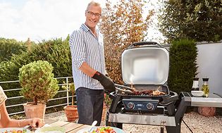 En man grillar på en elektrisk grill på en uteplats och tittar på en kvinna som sitter vid ett bord och ler mot mannen.