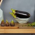 Bänkskiva i trä med fikon, en skål med aubergine och salt & pepparkvarn på. Mörkgrå vägg bakom. 