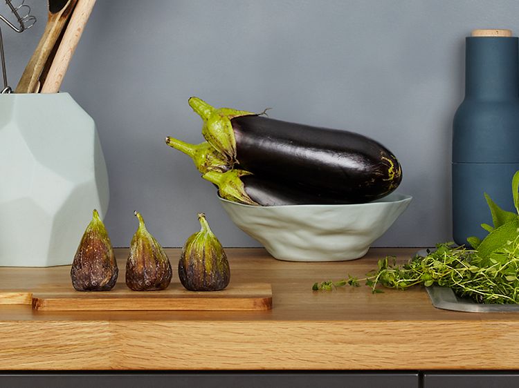 Bänkskiva i trä med fikon, en skål med aubergine och salt & pepparkvarn på. Mörkgrå vägg bakom.