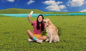 En tjej sitter på en grön gräsmatta bredvid en hund och tar en selfie med en OnePlus Nord 2-mobil. 