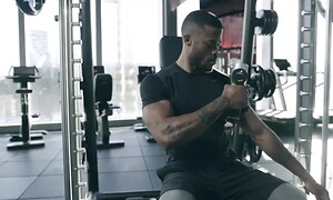 Man som sitter i en träningsmaskin på ett gym och använder en Hypervolt-massagepistol. 