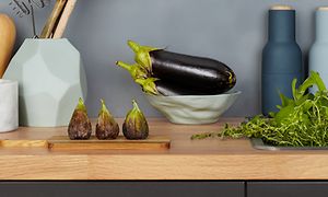 Närbild på en bänkskiva i trä med en skål med aubergine och fikon på en skärbräda. Salt och pepparkvarn syns i bakgrunden. 
