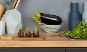 Närbild på en bänkskiva i trä med en skål med aubergine och fikon på en skärbräda. Salt och pepparkvarn syns i bakgrunden.