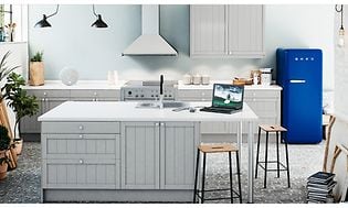 Ljust kök med en vit väggmonterad köksfläkt och ett blått kylskåp från Smeg och köksö med stolar. 