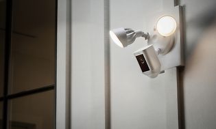 En vit övervakningskamera med lampor placerad utomhus på en vit vägg.