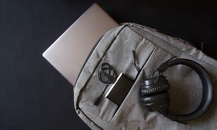 En grå ryggsäck med dator, hörlurar och powerbank som ligger ned på svart bakgrund.