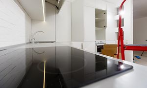 Keramikhäll installerad i kök där renovering pågår. Tomma hyllplan, kartonger och en röd stege syns i bakgrunden.