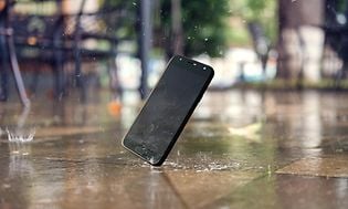 Mobiltelefon som ramlar ner på gatan och träffar asfalten så att skärmen krossas. Regndroppar faller och skvätter vatten. 