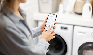 Kvinna som håller upp en mobiltelefon (med applikation som kan styra tvätten) framför en tvättmaskin i en tvättstuga.