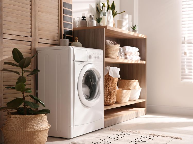 En liten tvättmaskin i en modern tvättstuga med en trähylla med olika saker på och en stor krukväxt bredvid. 