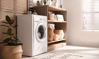 En liten tvättmaskin i en modern tvättstuga med en trähylla med olika saker på och en stor krukväxt bredvid. 