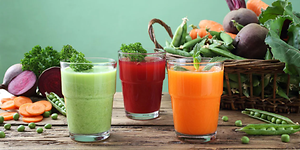 Tre glas med juice på ett bord med grönsaker. En grön juice, en orange och en röd. 