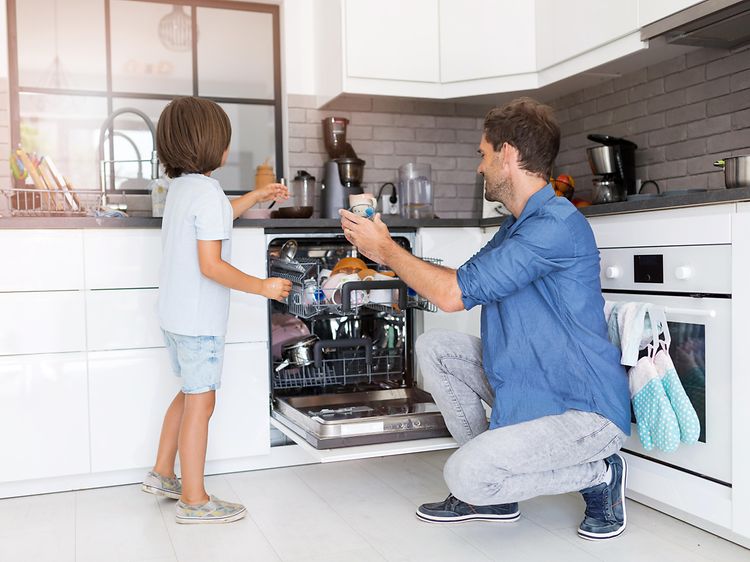 En liten pojke och en vuxen man plockar ur en liten diskmaskin tillsammans i ett vitt kök. Mannen står på knä. 