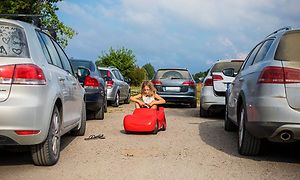 En flicka i en röd leksaksbil på en parkering med riktiga bilar runt omkring. 