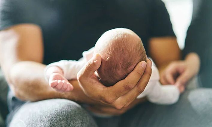 Man som håller en liten nyfödd baby i knäet med handen under huvudet.