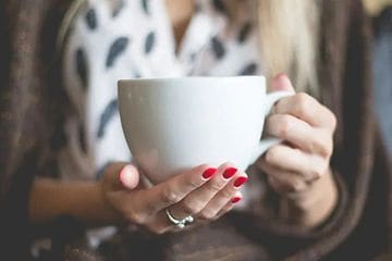 Närbild på en kvinna med röda naglar som håller i en stor vit kaffekopp. 