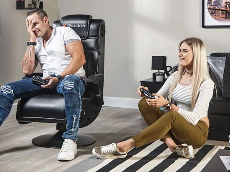 Man sitter i en svart X Rocker-stol och spelar på spelkonsol med en kvinna som sitter på golvet. 