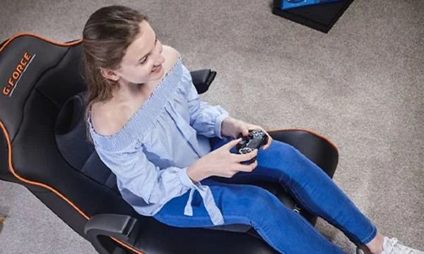 Kvinna som sitter bakåtlutad i en svart X Rocker-gamingstol med röda detaljer och spelar på en spelkonsol. 