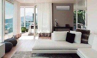 Vit värmepump upphängd på en brun vägg i ett ljust vardagsrum med stora fönster, en stor vit soffa och en platt-TV. 