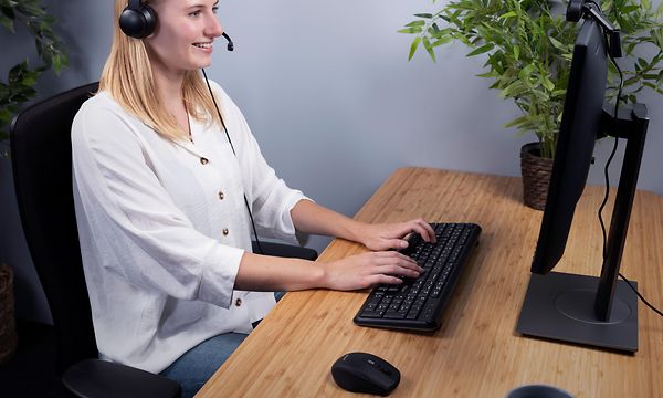Kvinna med headset på sig sitter och jobbar framför en dator på ett skrivbord i trä med en grön växt vid sidan. 