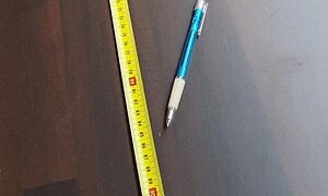 Penna och måttband som markerar en liten skada i en brun bänkskiva. Pennan visar var skadan är. 